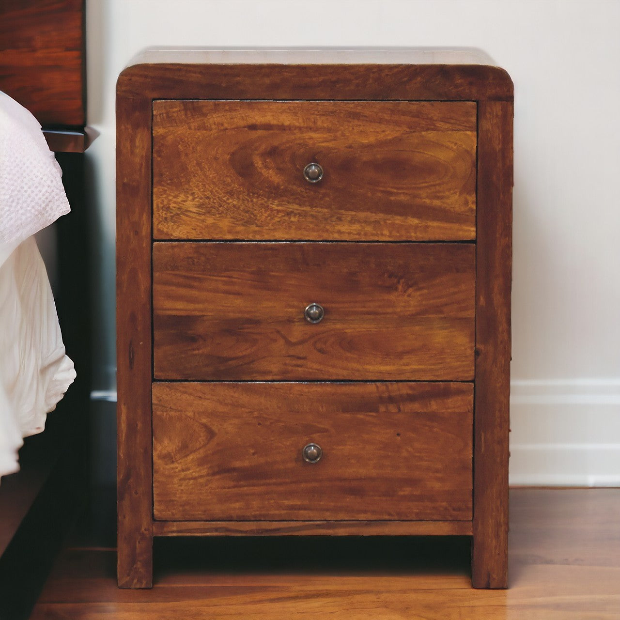 Naya Chestnut Nightstand made from solid mango wood with three drawers and brass knobs. Naya standing by a bedside.